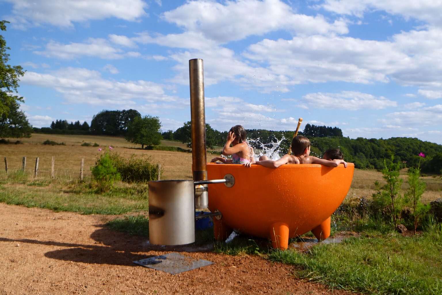 Drie kinderen spetteren in een oranje Dutchtub, met de houtkachel ernaast en weilanden op de achtergrond